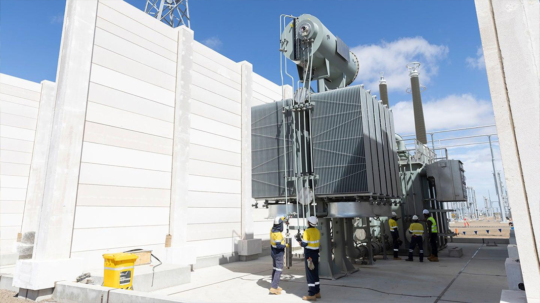 Workers beside a power transformer and radiator bank between outdoor concrete walls